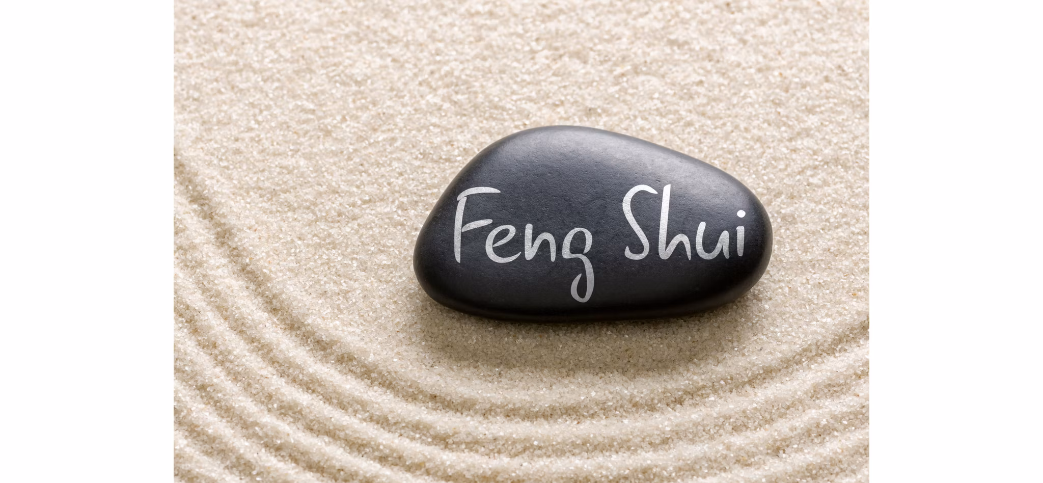 A smooth black stone with 'Feng Shui' written on it, resting on a patterned sand background.