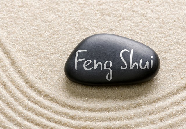 A smooth black stone with 'Feng Shui' written on it, resting on a patterned sand background.