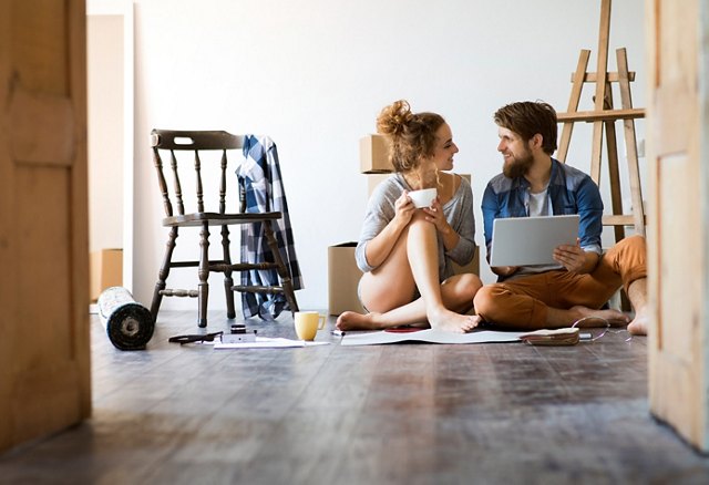 A couple sitting on the floor, looking at a laptop and enjoying coffee among moving boxes and furniture.