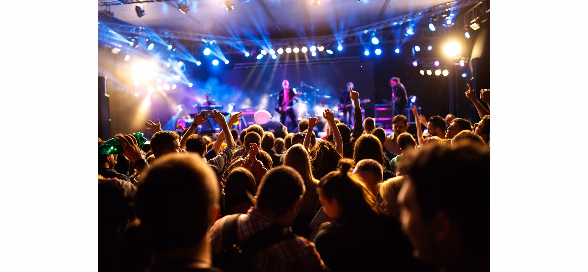 Energetic crowd enjoying a live concert with hands raised and a band performing on stage under vibrant stage lights.