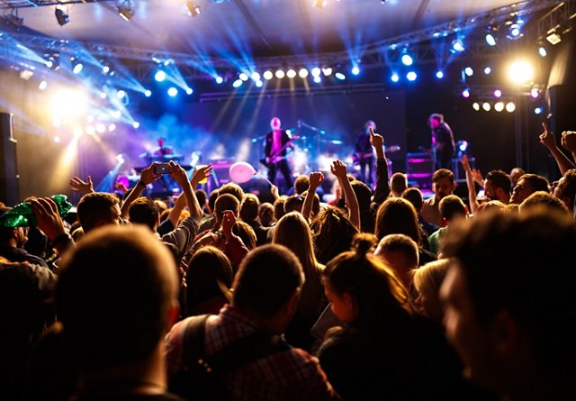 Energetic crowd enjoying a live concert with hands raised and a band performing on stage under vibrant stage lights.