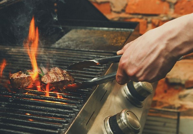 A person grilling steaks over an open flame on an outdoor barbecue.