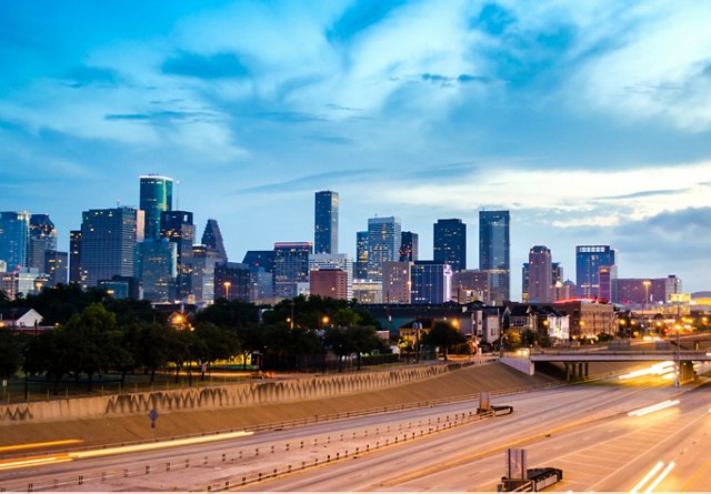 Twilight skyline of Houston with illuminated buildings and a clear highway in the foreground.