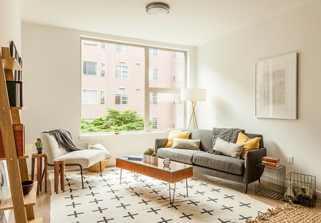 Cozy living room with a large window, gray sofa with yellow accents, patterned rug, and a tall floor lamp.