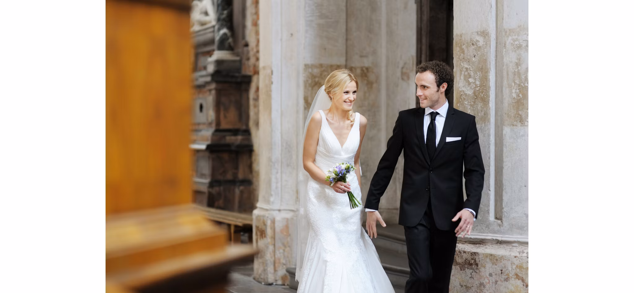 A bride and groom walking hand in hand, smiling at each other, in a historical building.