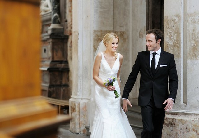 A bride and groom walking hand in hand, smiling at each other, in a historical building.