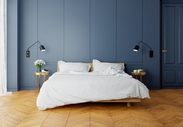 Modern minimalist bedroom with an unmade bed, wooden side tables, and wall-mounted lamps against a dark blue wall.