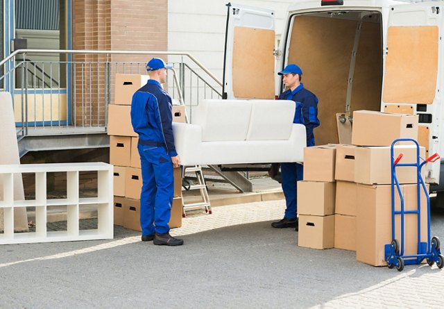 Two movers in blue uniforms loading a white sofa and cardboard boxes into a moving van.