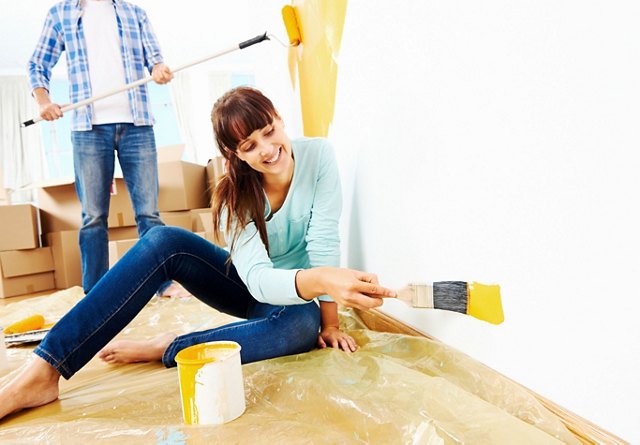 A woman sitting on the floor painting a wall yellow with a man behind her using a roller, both surrounded by painting supplies and cardboard boxes.