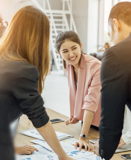 A team of professionals gathered around a table discussing charts and data, with a focus on the smiling woman in a pink blazer engaging her colleagues.