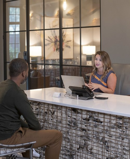 A man and a woman engaging in conversation across a reception desk, with the woman attending to a computer, in an office with modern decor.