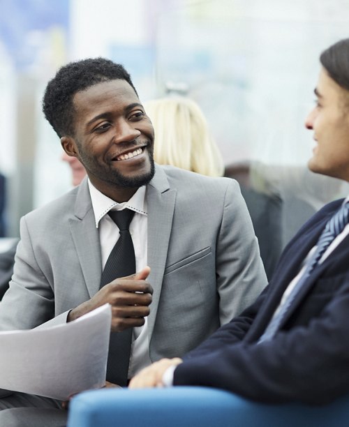 A cheerful businessman in a gray suit holding documents and engaging in a discussion with a colleague, in a modern office setting.