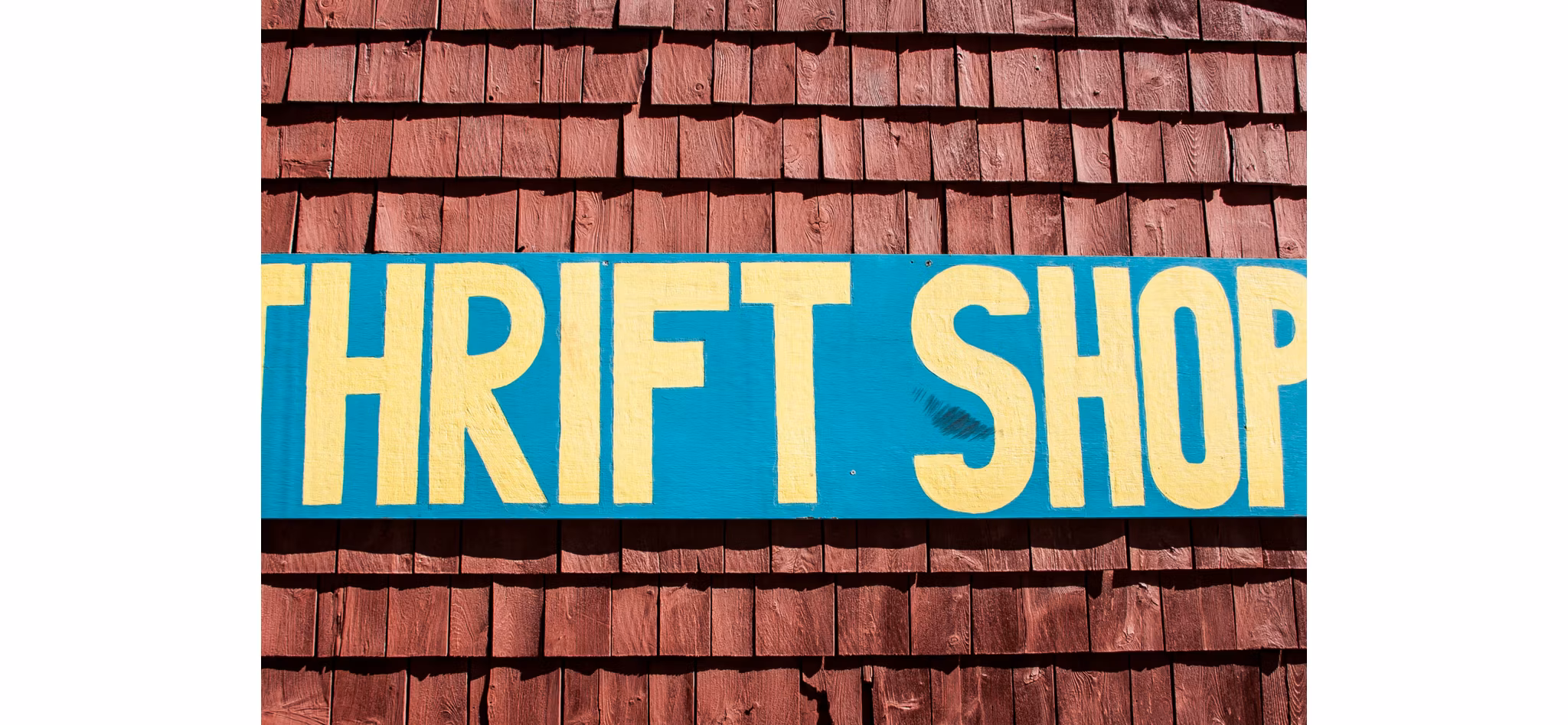A colorful sign with the words 'THRIFT SHOP' painted in bold yellow letters on a teal background, mounted on a wooden shingle wall.