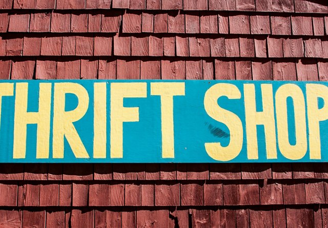 A colorful sign with the words 'THRIFT SHOP' painted in bold yellow letters on a teal background, mounted on a wooden shingle wall.