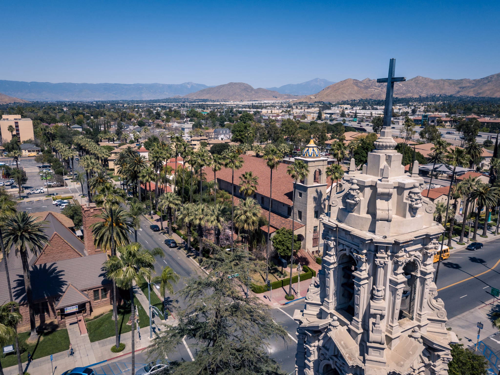 A historic church tower with a cross overlooks palm-lined streets and distant mountains in Riverside, California, under a clear blue sky.