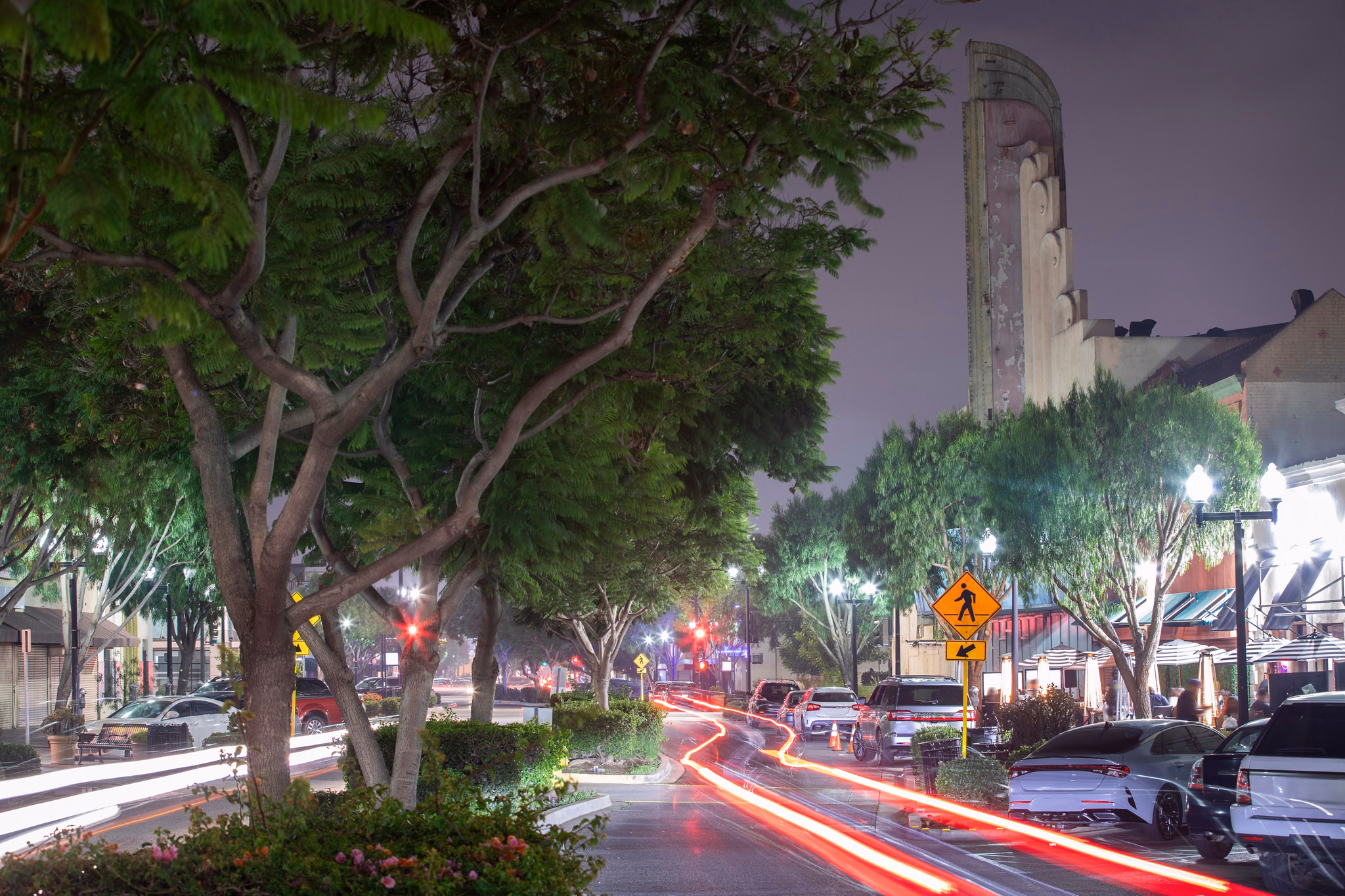 A lively street in Inglewood, California, at night with glowing streetlights, trees lining the road, and light trails from passing cars.