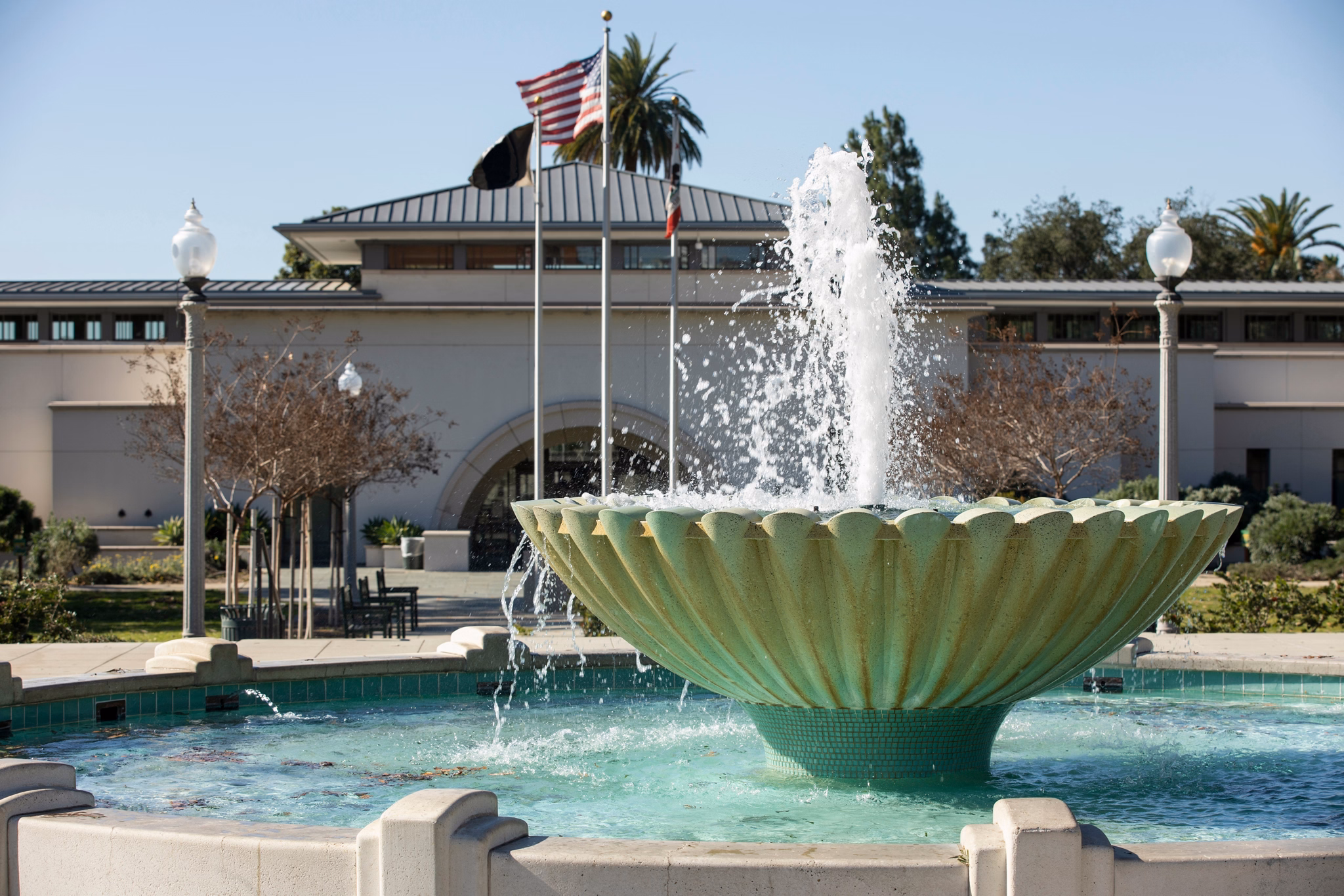 Fountain in Monrovia, California, with a flowing water feature, flagpoles, and a civic building framed by trees in the background.