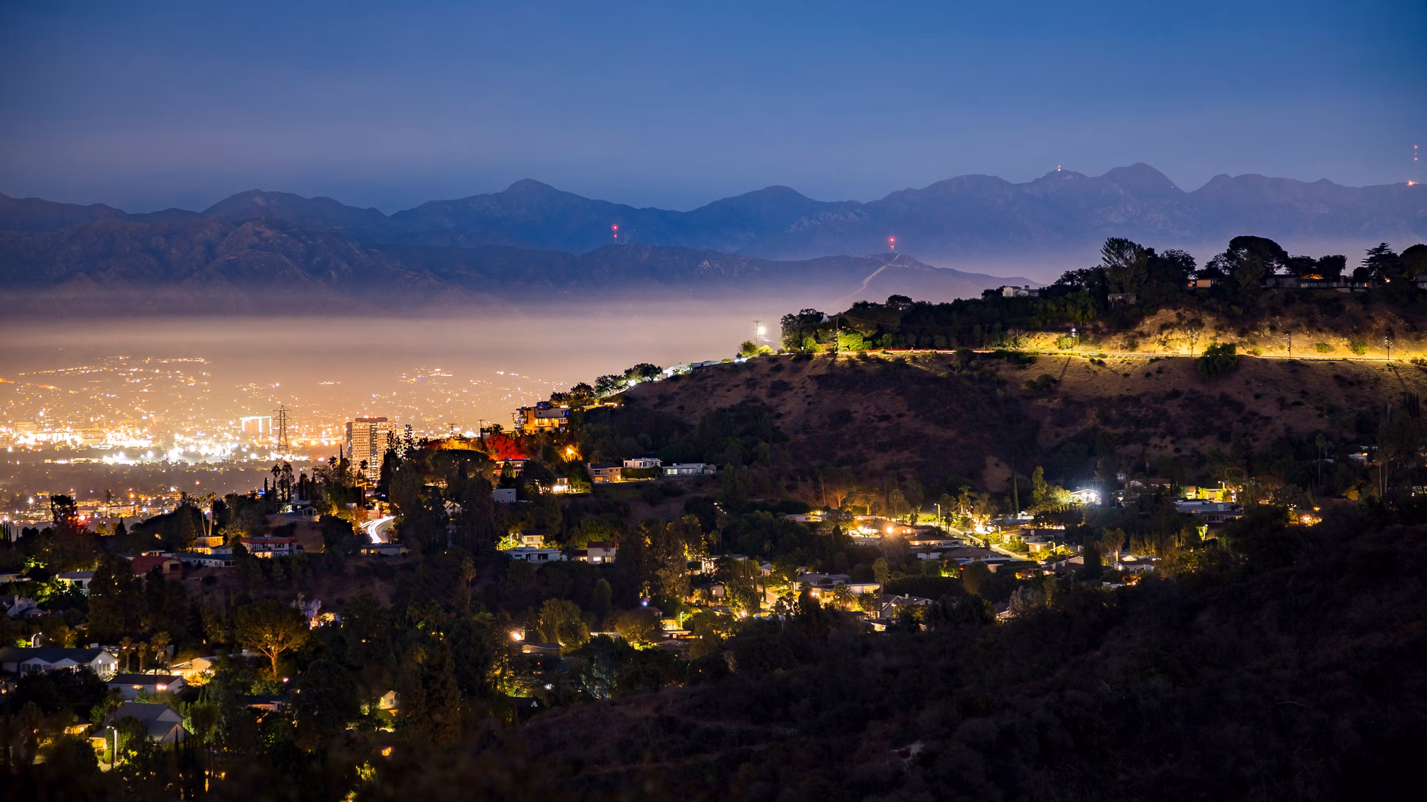 A nighttime view of North Hollywood, California, with glowing city lights, hillside homes, and mountains in the background under a dark sky.