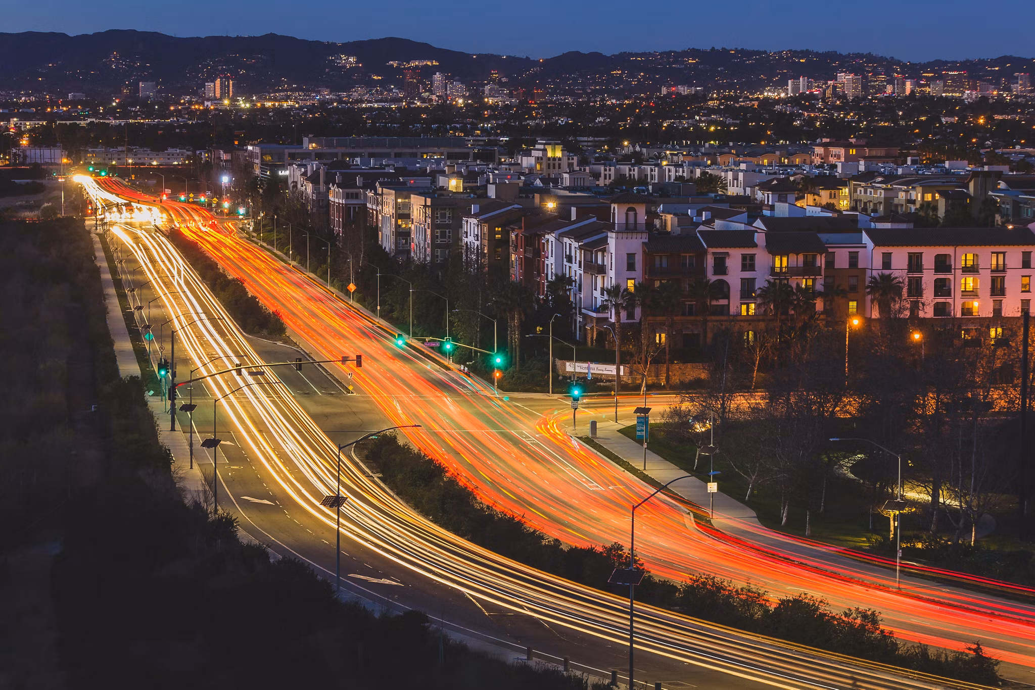 A vibrant night view of traffic lights streaking along a busy road in Playa Vista, California, with city lights and hills glowing in the background.