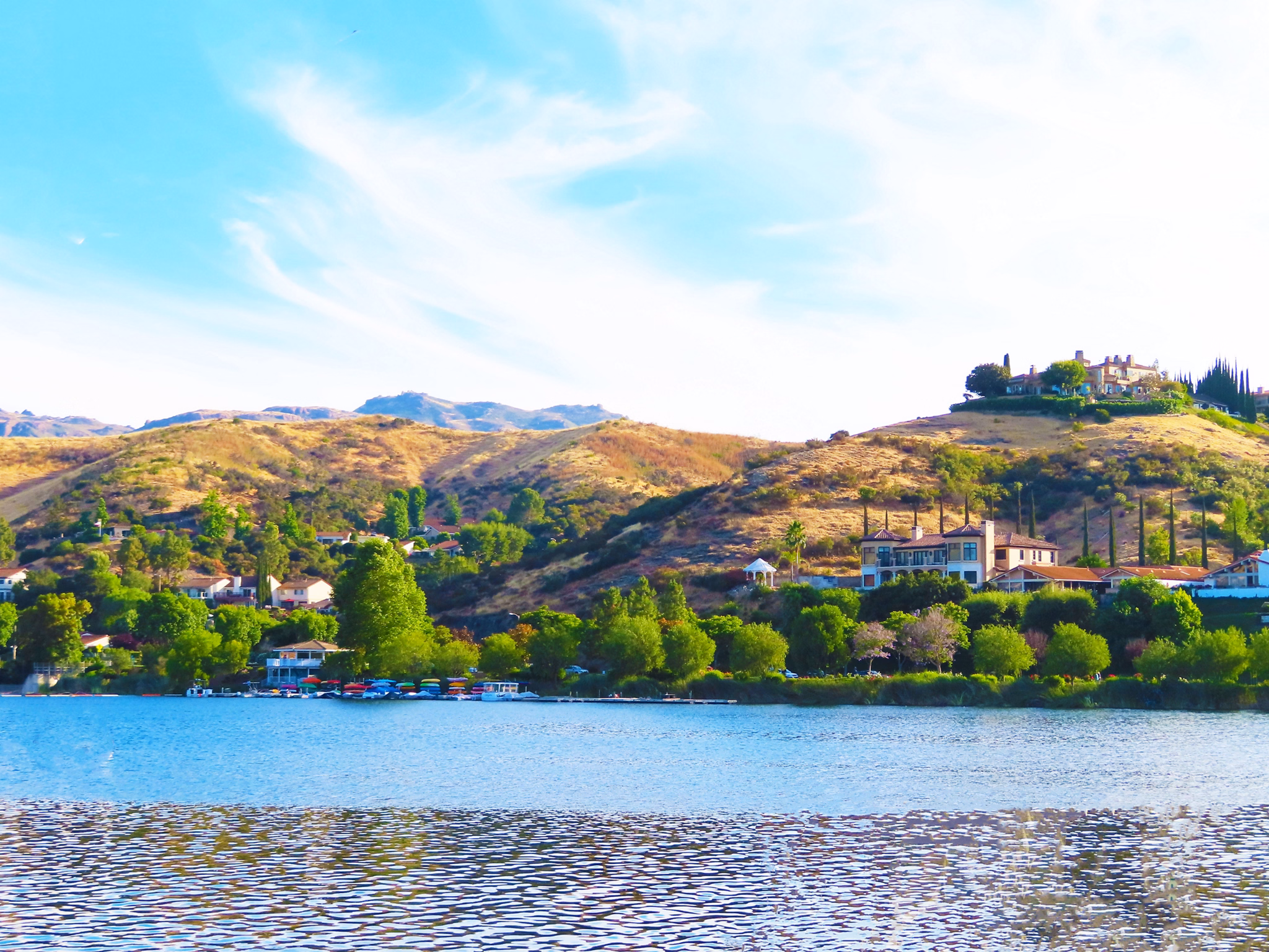 A peaceful lake with hillside homes surrounded by greenery in Westlake, California, under a bright blue sky with wispy clouds.