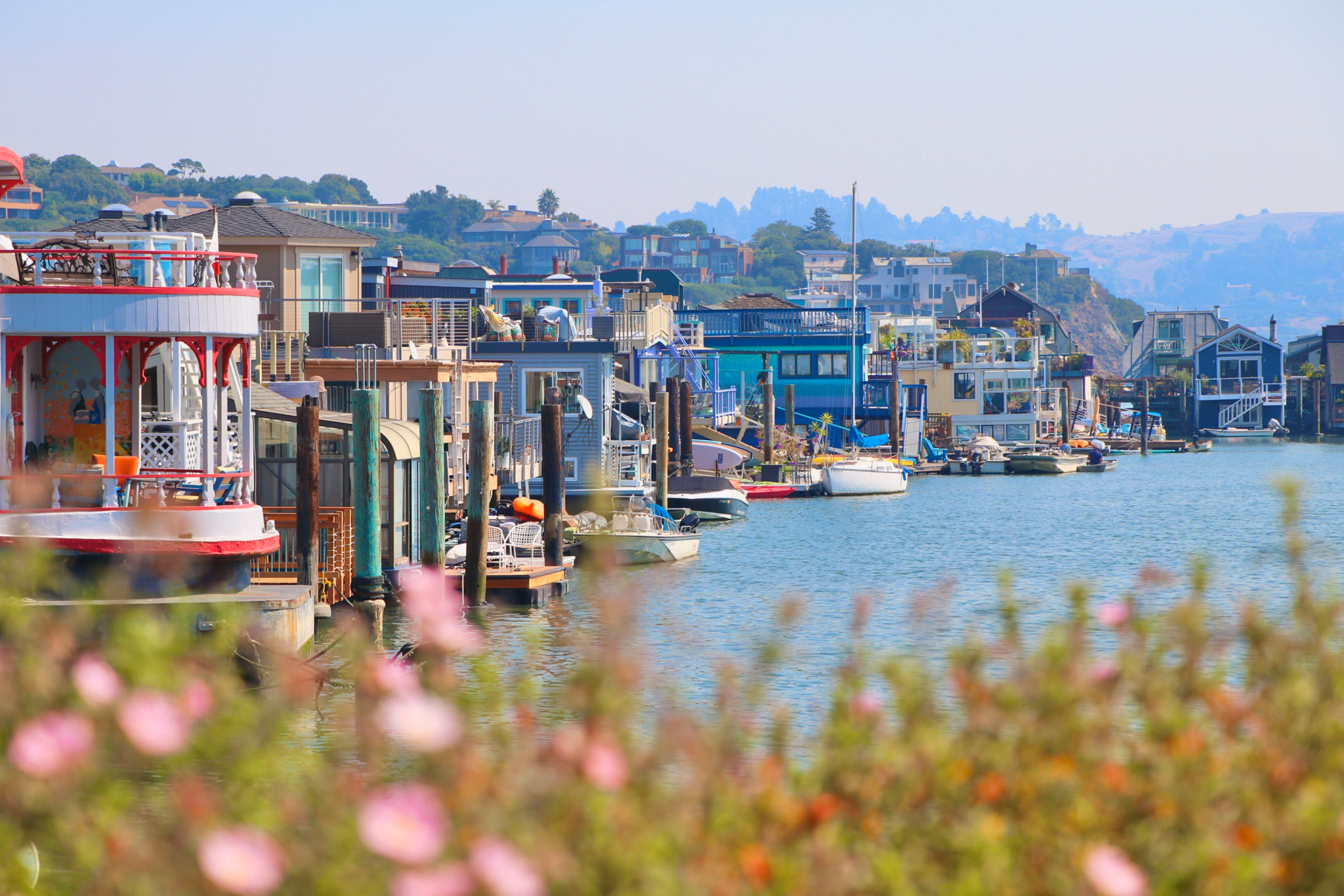 Colorful houseboats line a calm waterfront in Larkspur, California, with boats docked and hills in the background under a hazy sky.