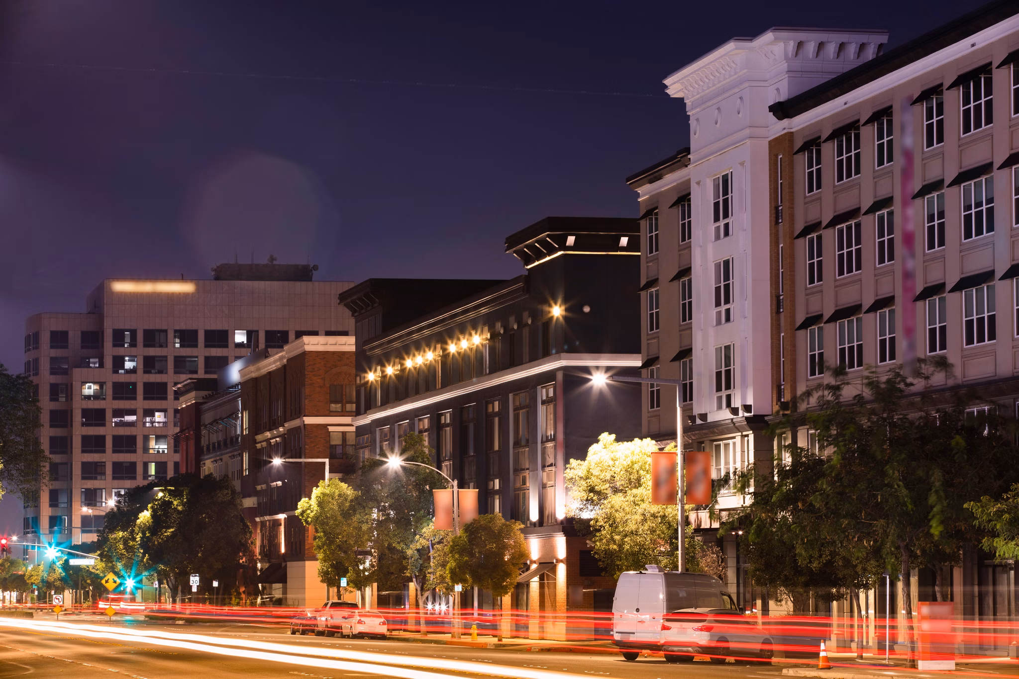 A nighttime view of lit-up buildings along a street in Anaheim, California, with streaks of car lights and trees lining the road.