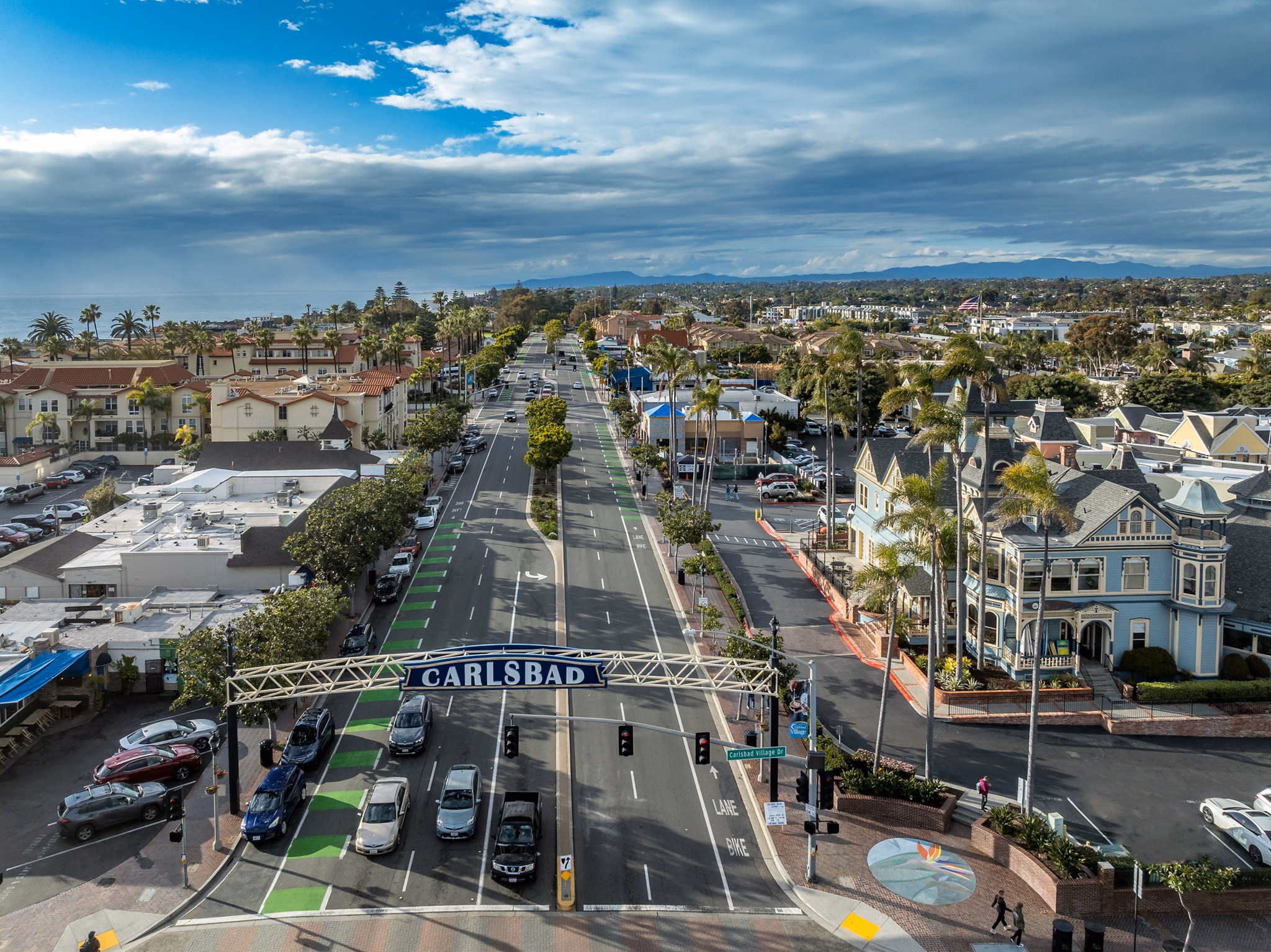 Aerial view of Carlsbad, California, featuring a tree-lined street, historic buildings, and a sign reading &quot;Carlsbad&quot; under a partly cloudy sky.