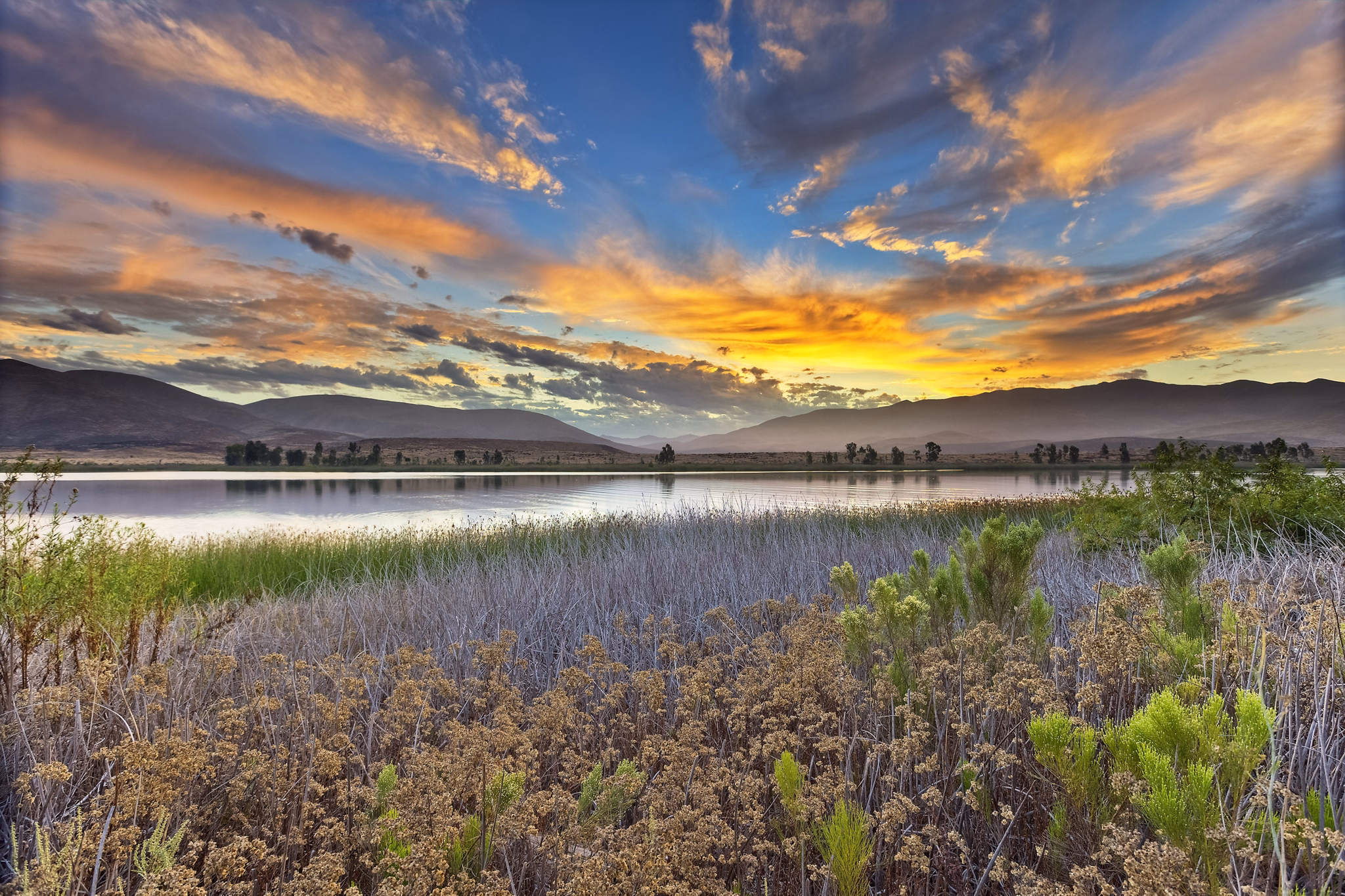 Scenic view of a serene lake at sunset with colorful skies, mountains, and lush vegetation in Otay Ranch, California.