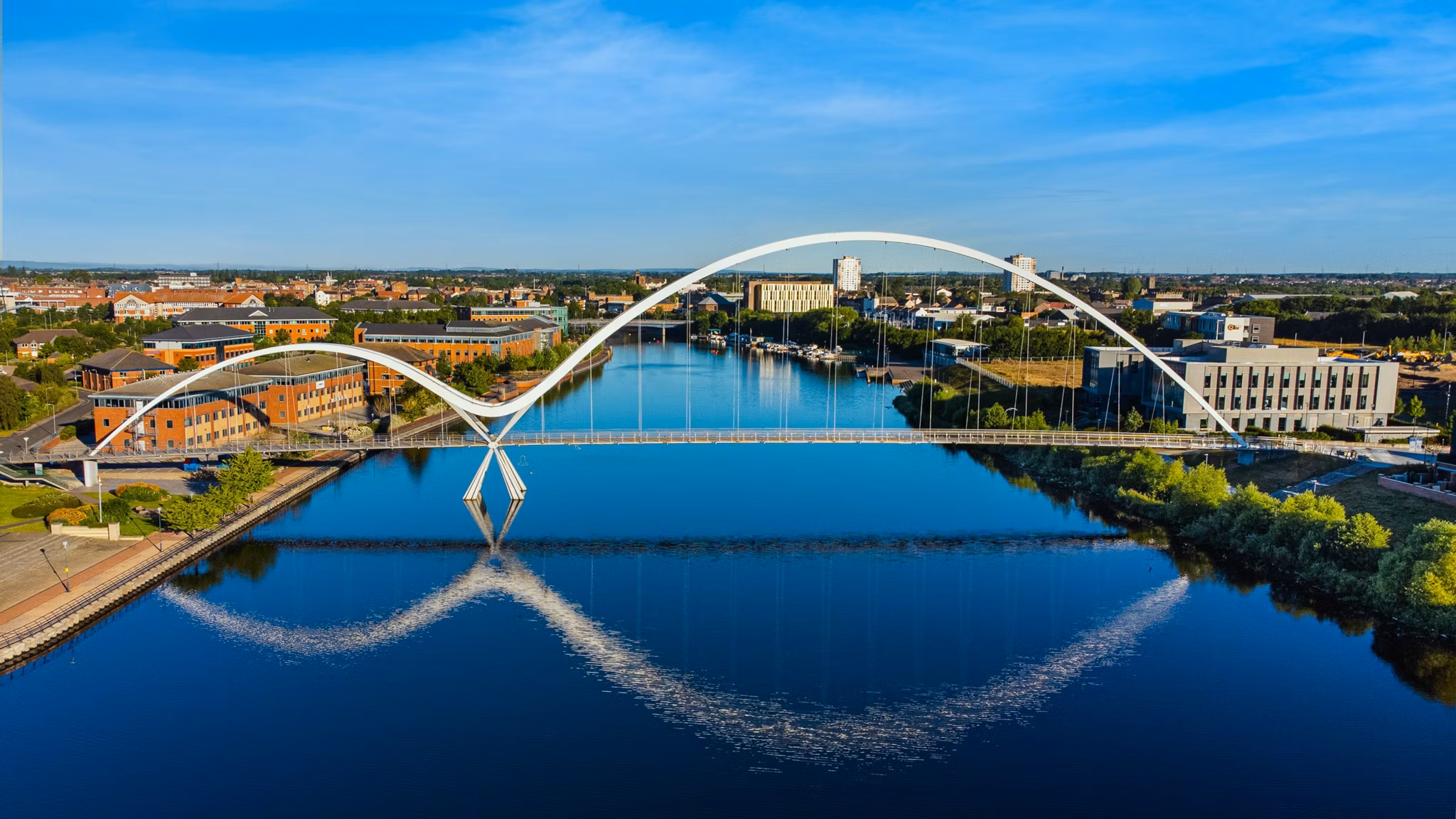 A modern arched bridge reflecting in a calm river, surrounded by buildings and greenery in Stockton, California, under a clear blue sky.