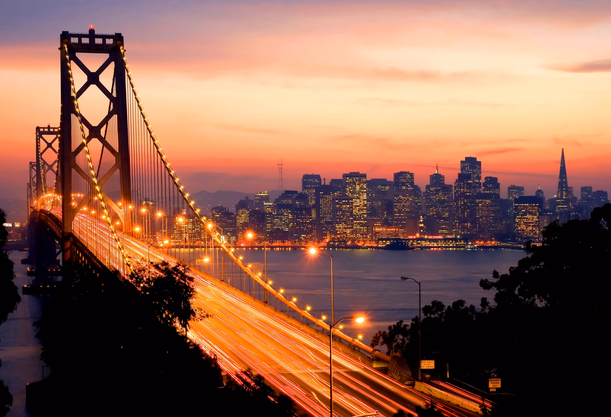 Bay Bridge glows with car lights at sunset, connecting to San Francisco, California&apos;s illuminated downtown skyline.