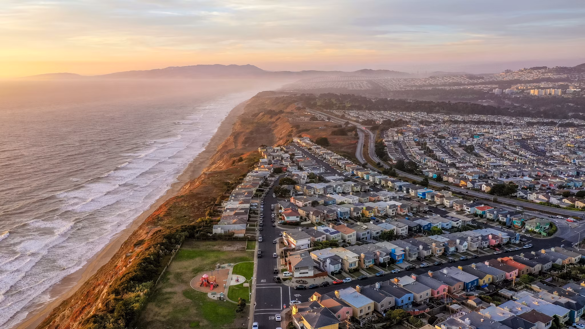 Sunset aerial view of Daly City, California, with colorful houses along the coastline, ocean waves, and misty cliffs at dusk.