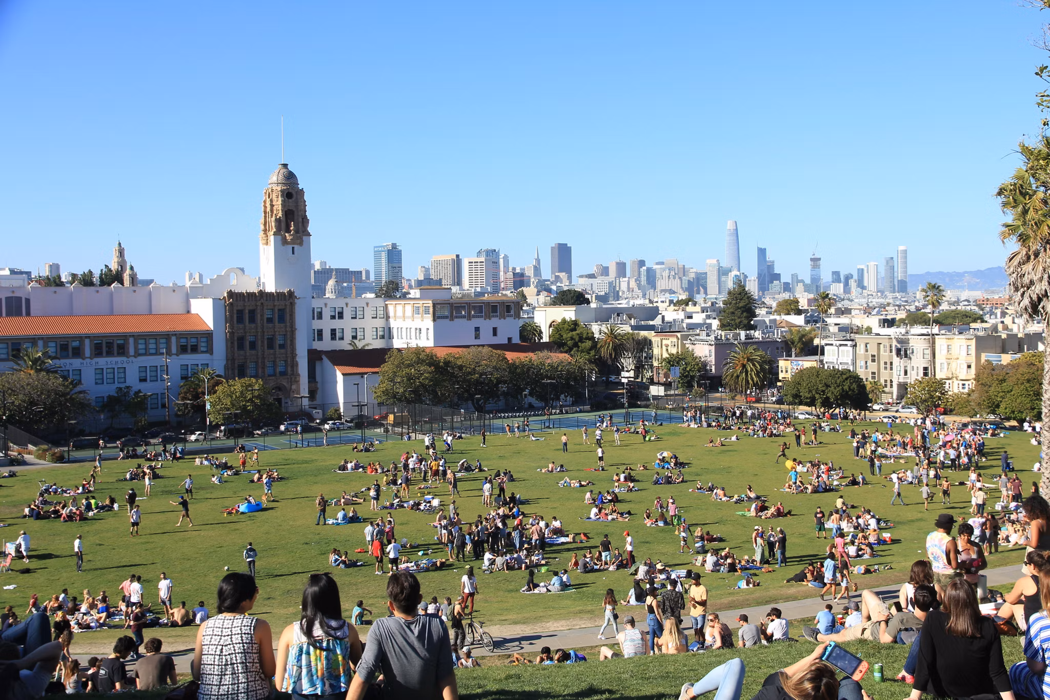 Crowded grassy park with city skyline, including tower building, in Mission District, California.
