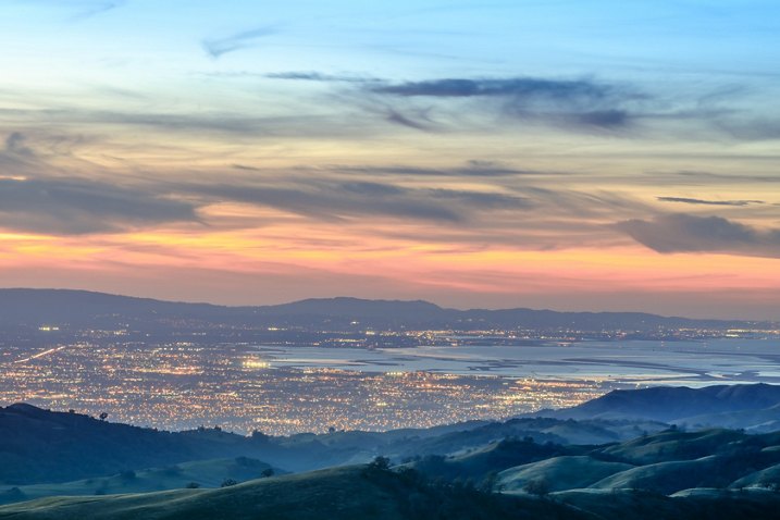 Sunset view of Santa Clara, California, from rolling hills, with city lights twinkling across the valley and bay below.