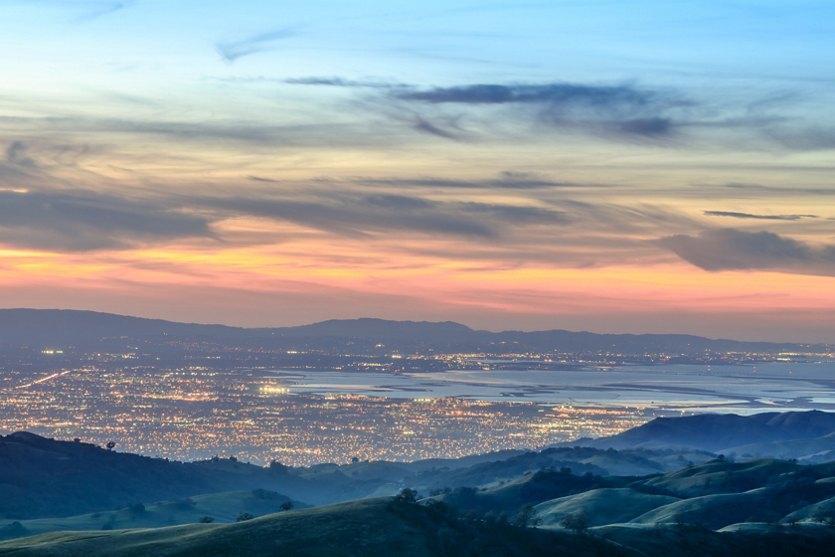 Sunset view of Santa Clara, California, from rolling hills, with city lights twinkling across the valley and bay below.