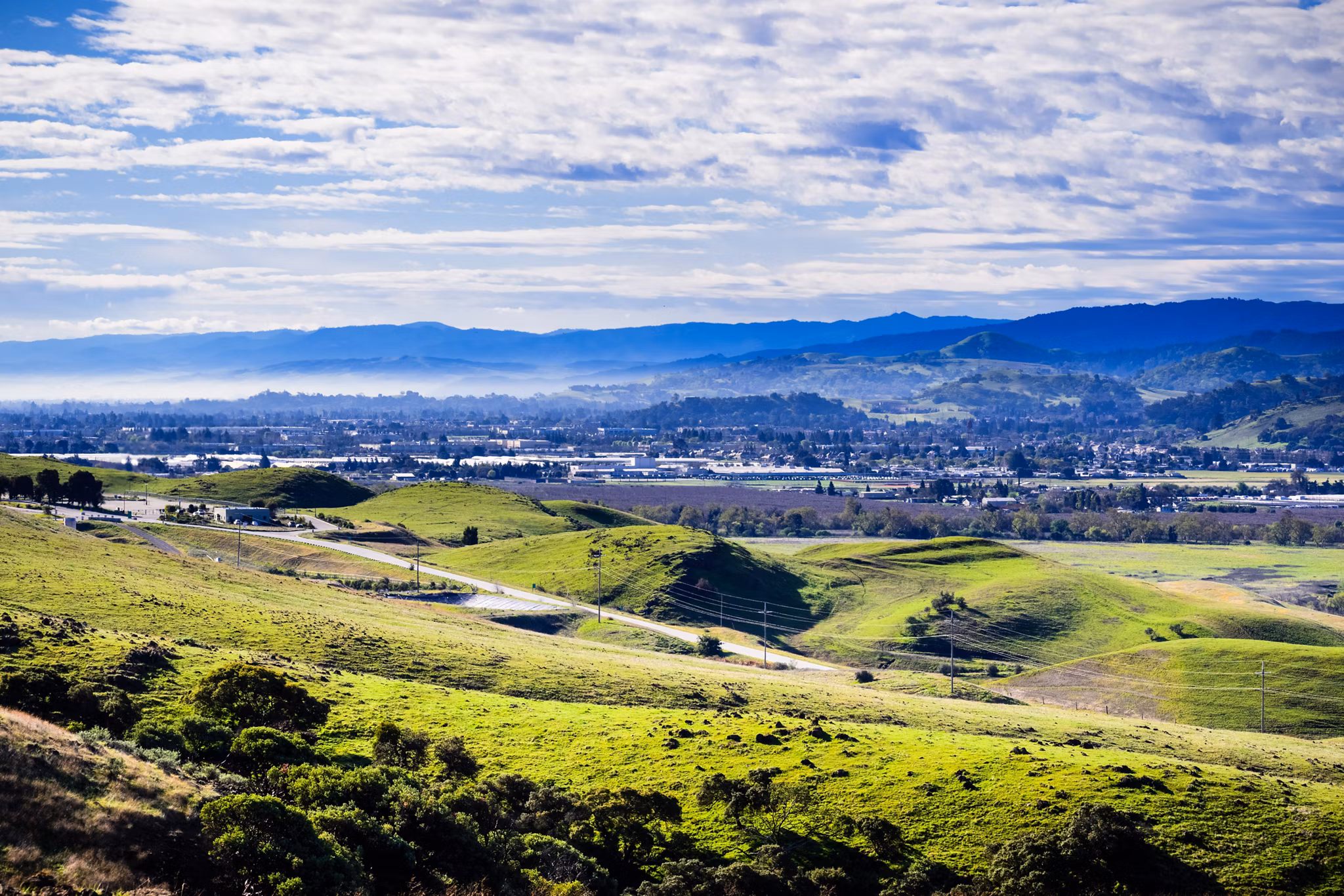 Rolling green hills overlook the valley town of Morgan Hill, California, with distant mountains and scattered clouds in a blue sky.