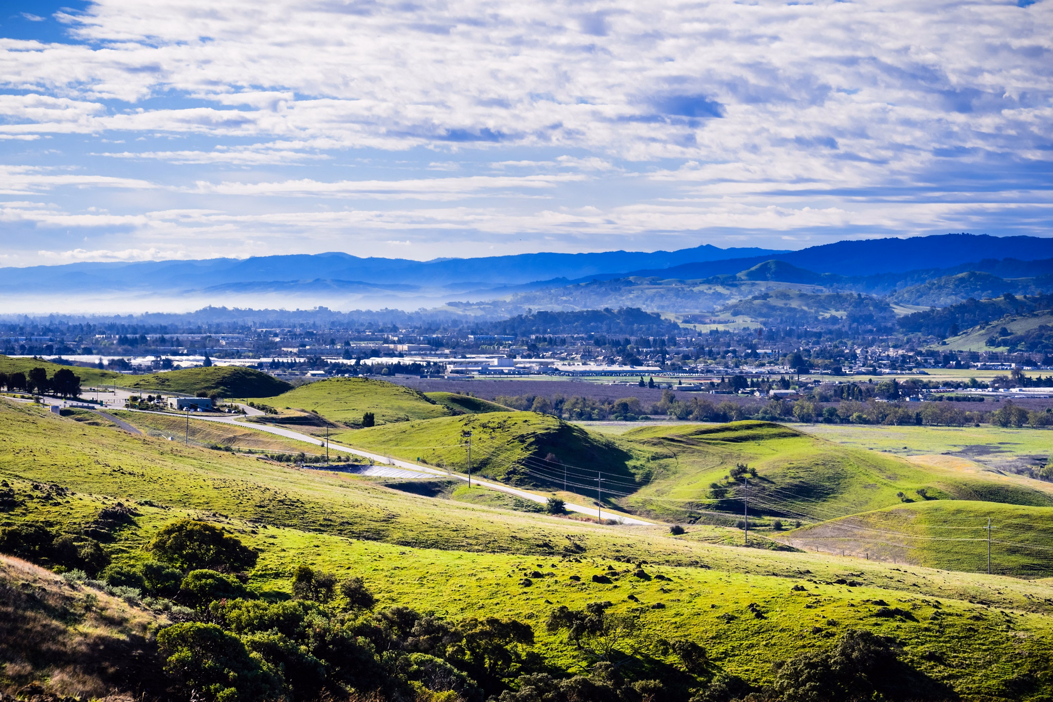 Rolling green hills overlook the valley town of Morgan Hill, California, with distant mountains and scattered clouds in a blue sky.