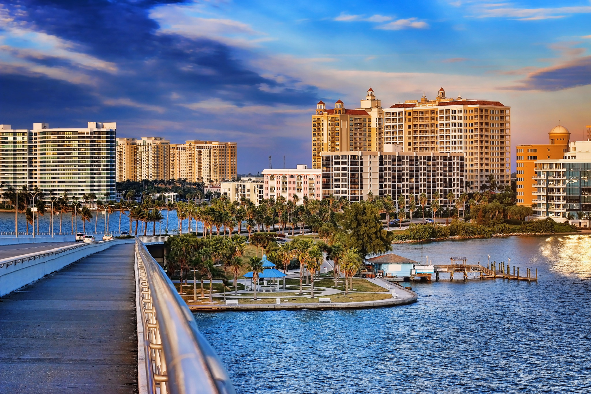 View of Sarasota, Florida, showing luxury waterfront condos, palm tree-lined park, and blue water, seen from a bridge.