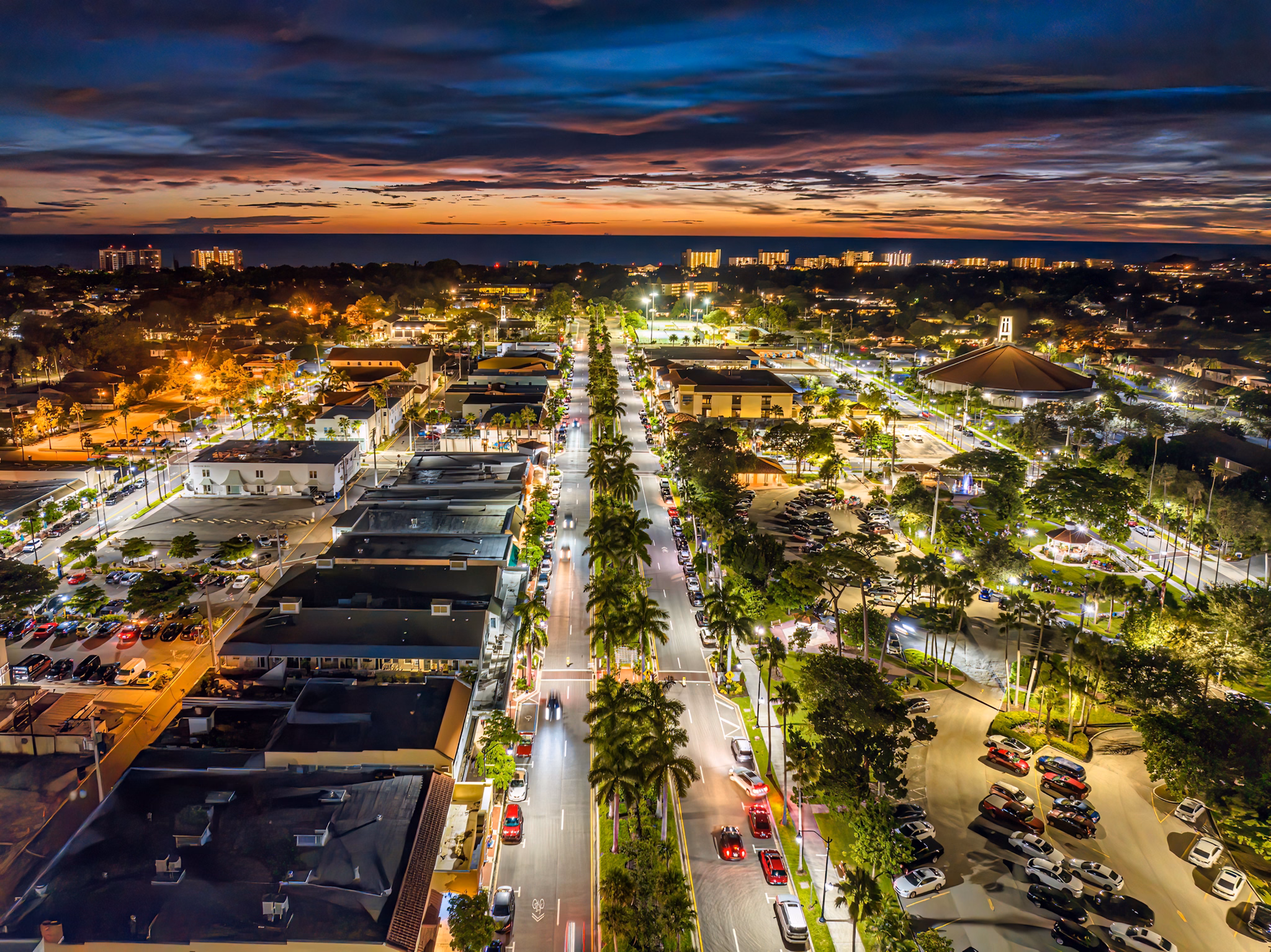 Night view of Venice, Florida, with a palm-lined main street leading to the ocean, colorful sunset sky, and illuminated buildings.
