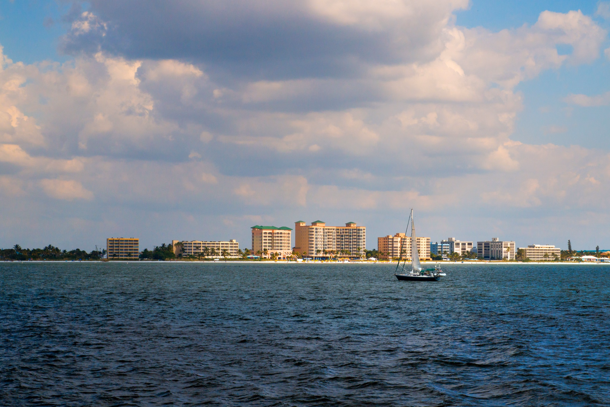 View of Estero, Florida&apos;s beachfront with a sailboat on dark blue water and beachside condos under puffy clouds.