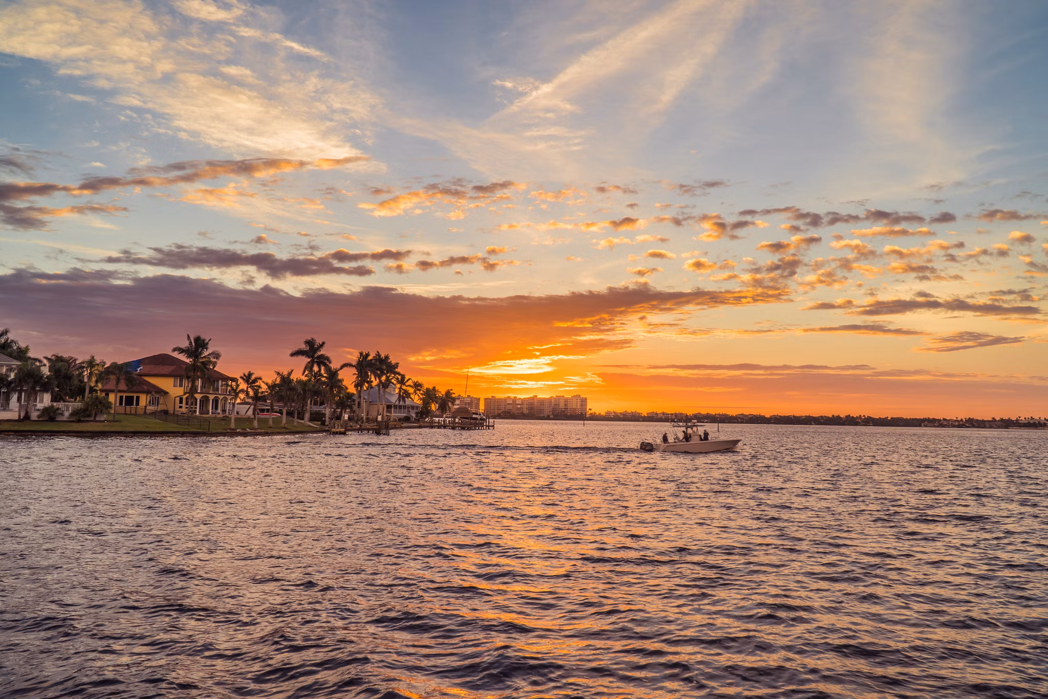 Stunning sunset over waterfront in Cape Coral, Florida, with a small boat cruising past waterfront homes with palm trees and colorful sky reflecting on the water.