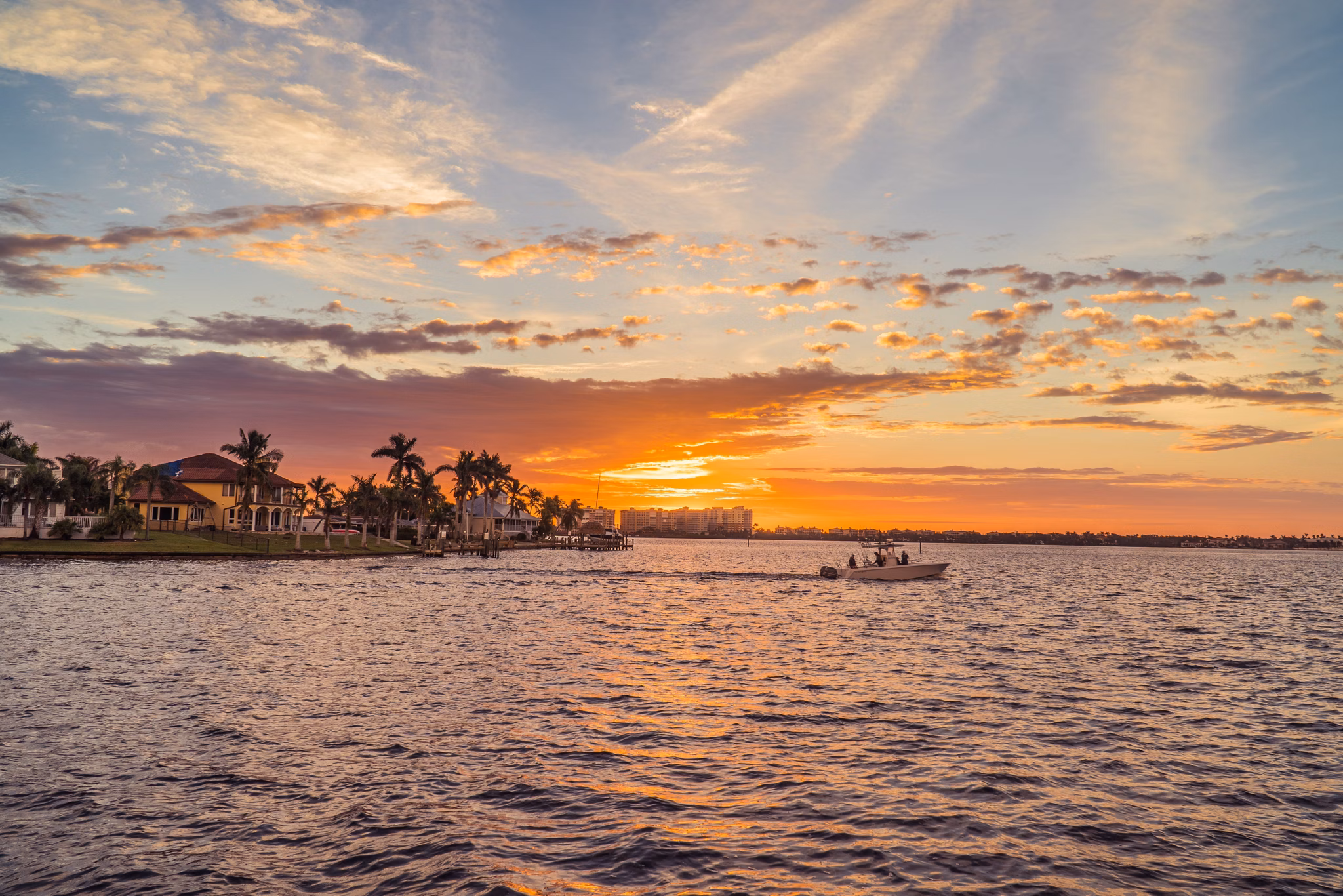 Stunning sunset over waterfront in Cape Coral, Florida, with a small boat cruising past waterfront homes with palm trees and colorful sky reflecting on the water.