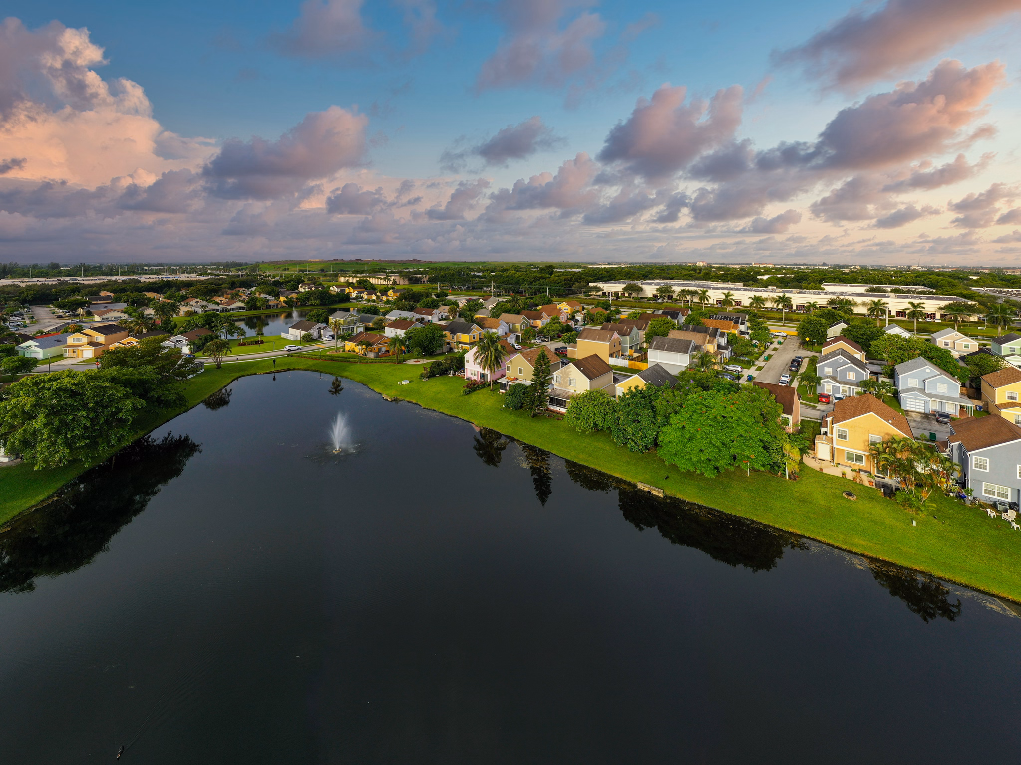 Aerial view of Miramar/Hialeah, Florida, showing colorful residential homes surrounding a retention pond with water fountain under pink clouds.