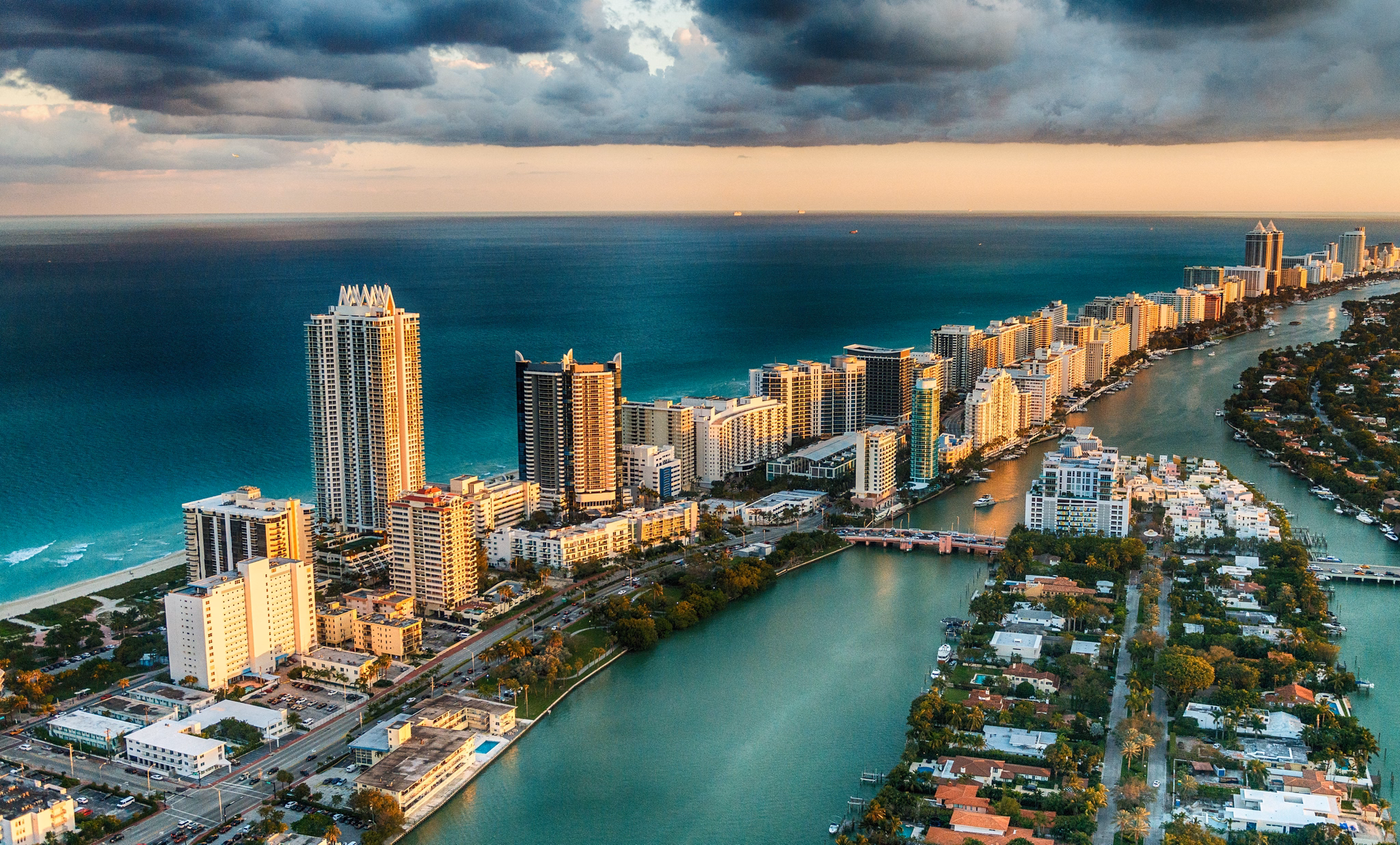 Aerial view of South Miami, Florida, with high-rise condos along the ocean, waterways separating beachfront from residential areas, under dramatic storm clouds.