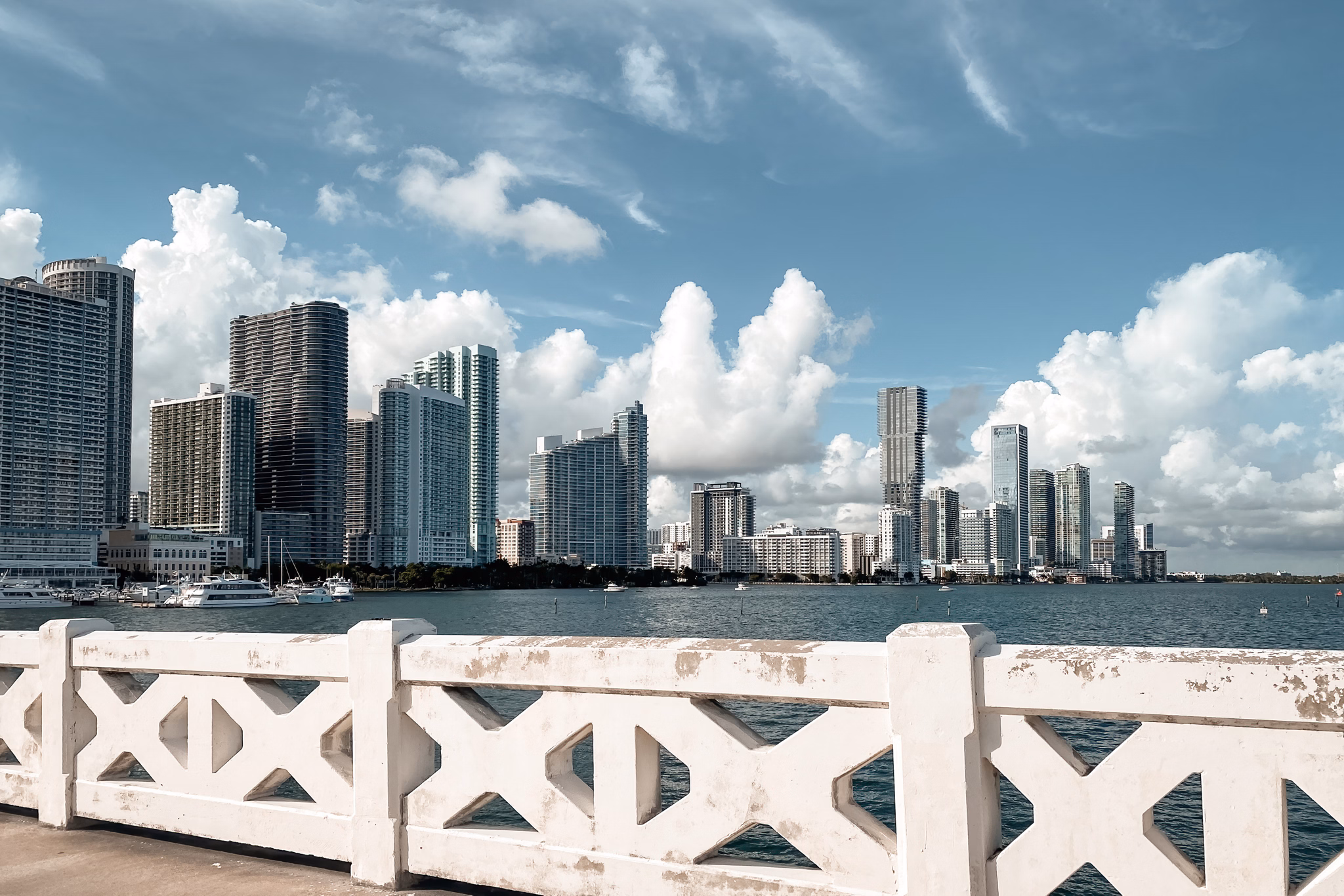 View of Miami Design District, Florida skyline across Biscayne Bay, with tall waterfront condos and office buildings under fluffy clouds, seen from behind a white railing.