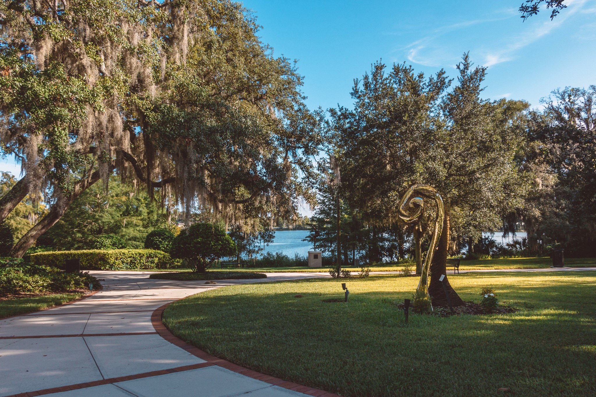 Park pathway in Seminole County, Florida, with Spanish moss hanging from oak trees, lawn sculpture, and blue lake in background.