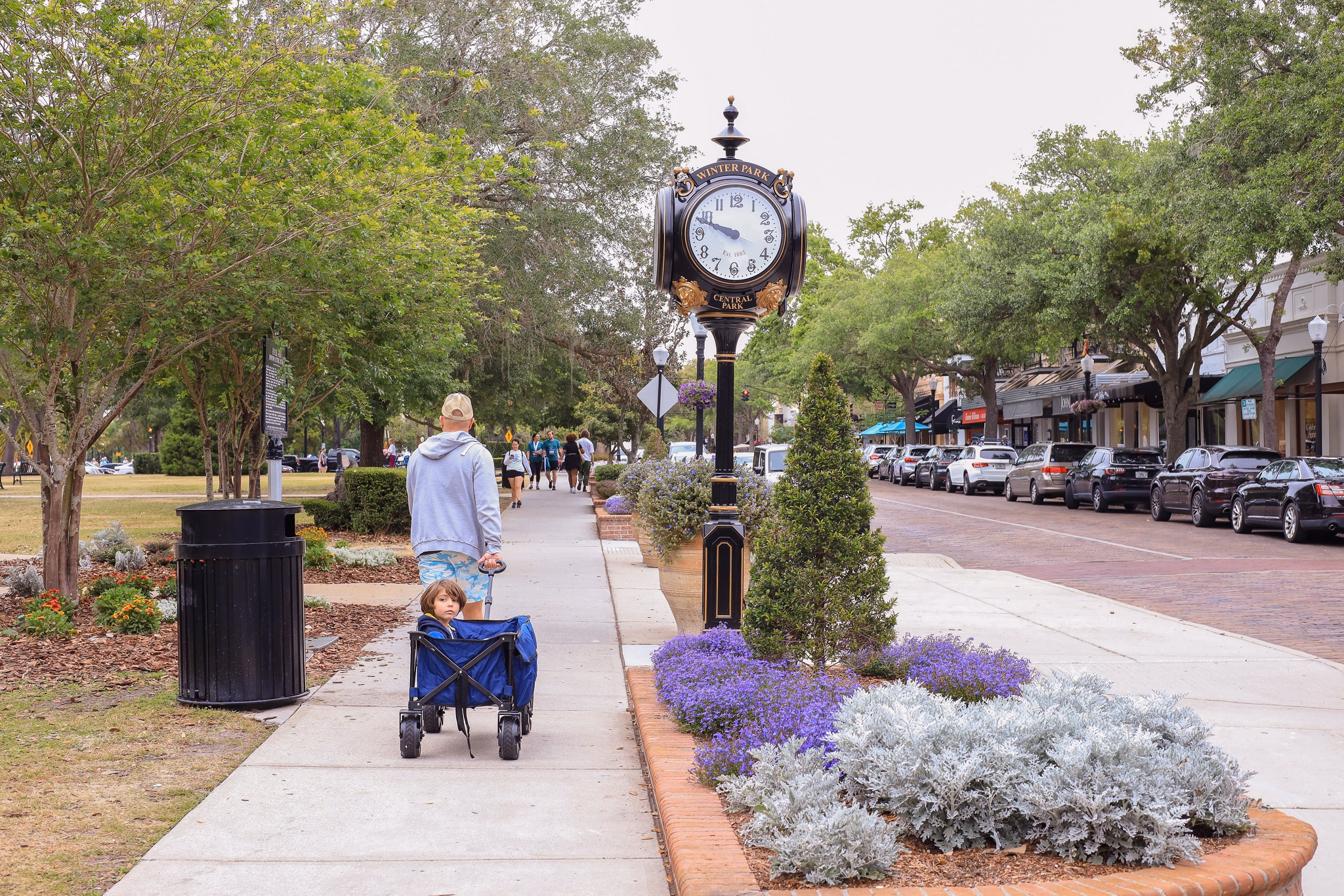 Downtown Winter Park, Florida&apos;s tree-lined sidewalk with decorative clock, purple flowers, and people walking past shops.