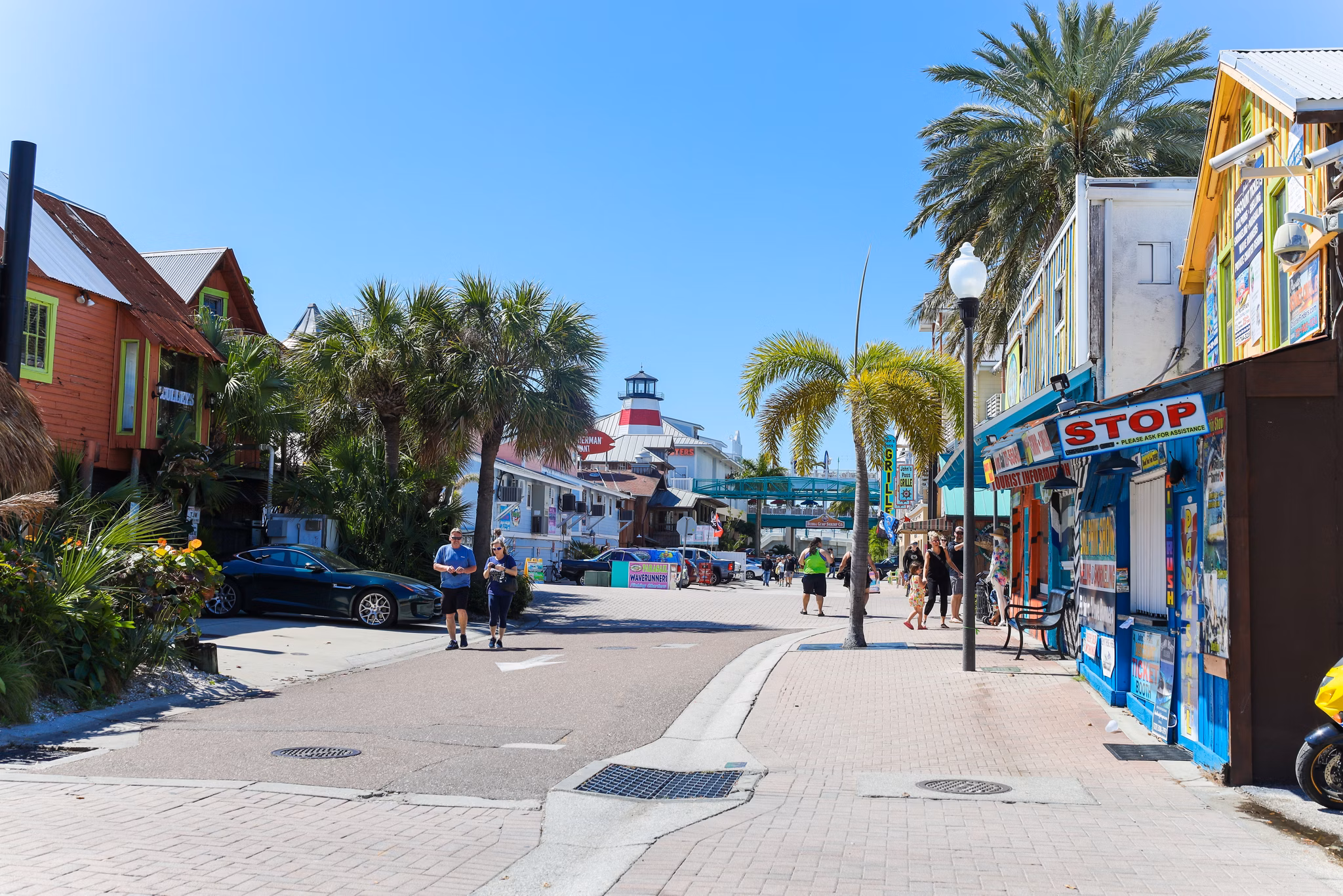 Colorful shopping street in Pinellas County, Florida, with lighthouse structure visible, palm trees, and tourists walking between shops.