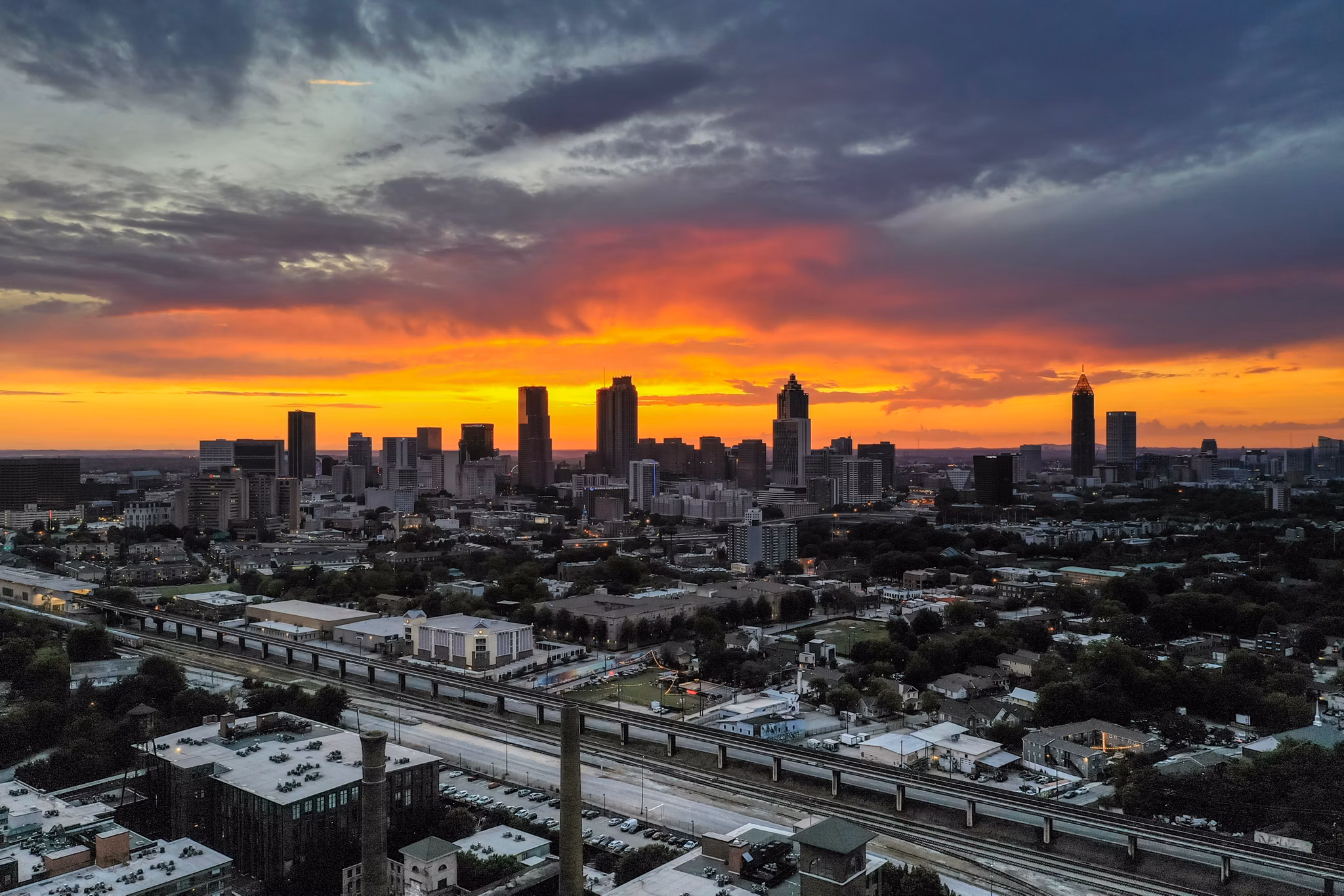 Dramatic sunset over Atlanta skyline viewed from Grant Park, GA with orange-red sky, silhouetted skyscrapers, and transit lines below.