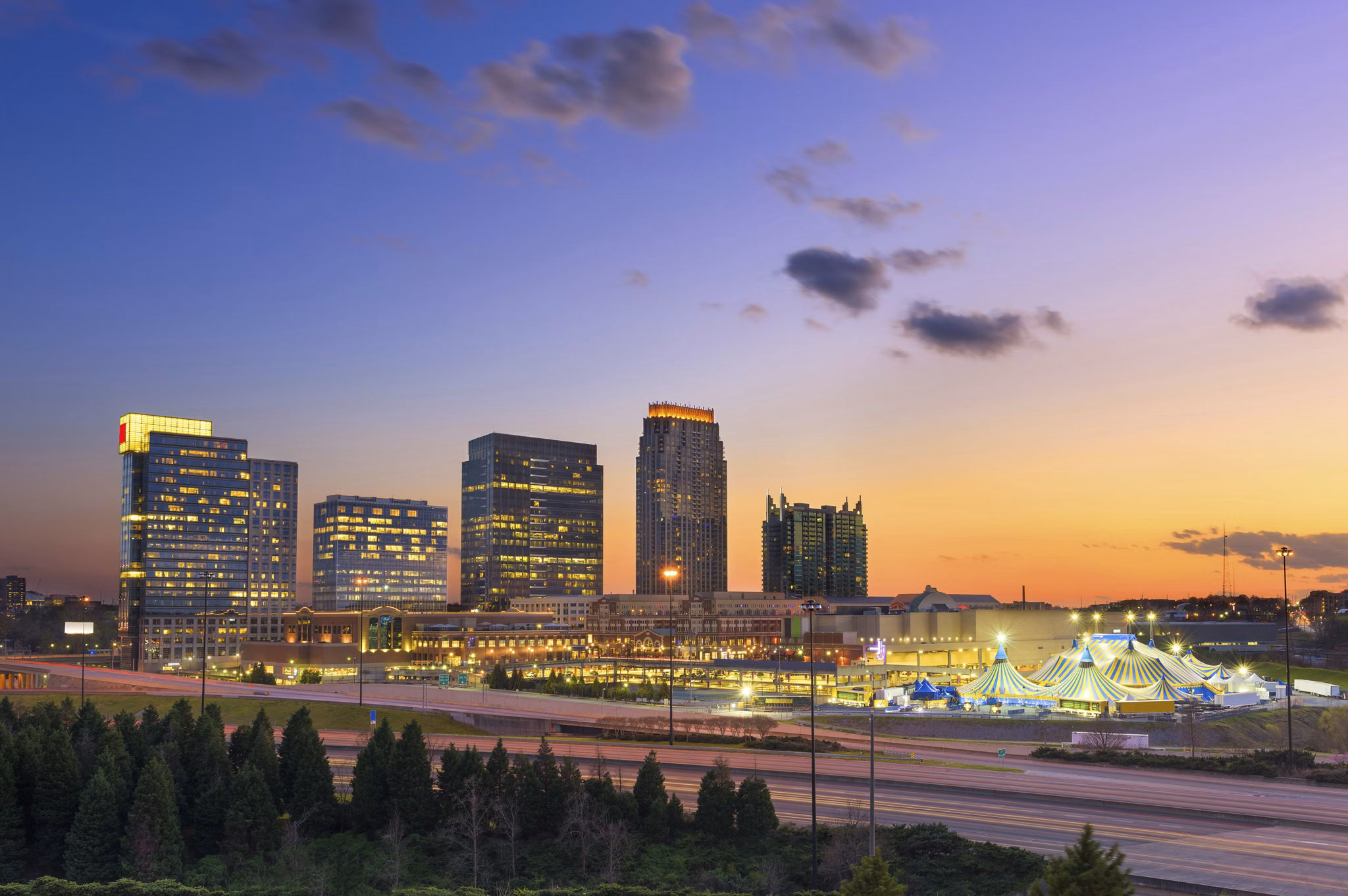 Marietta, GA at sunset with illuminated high-rise buildings, highway with light trails, and blue circus tent below.