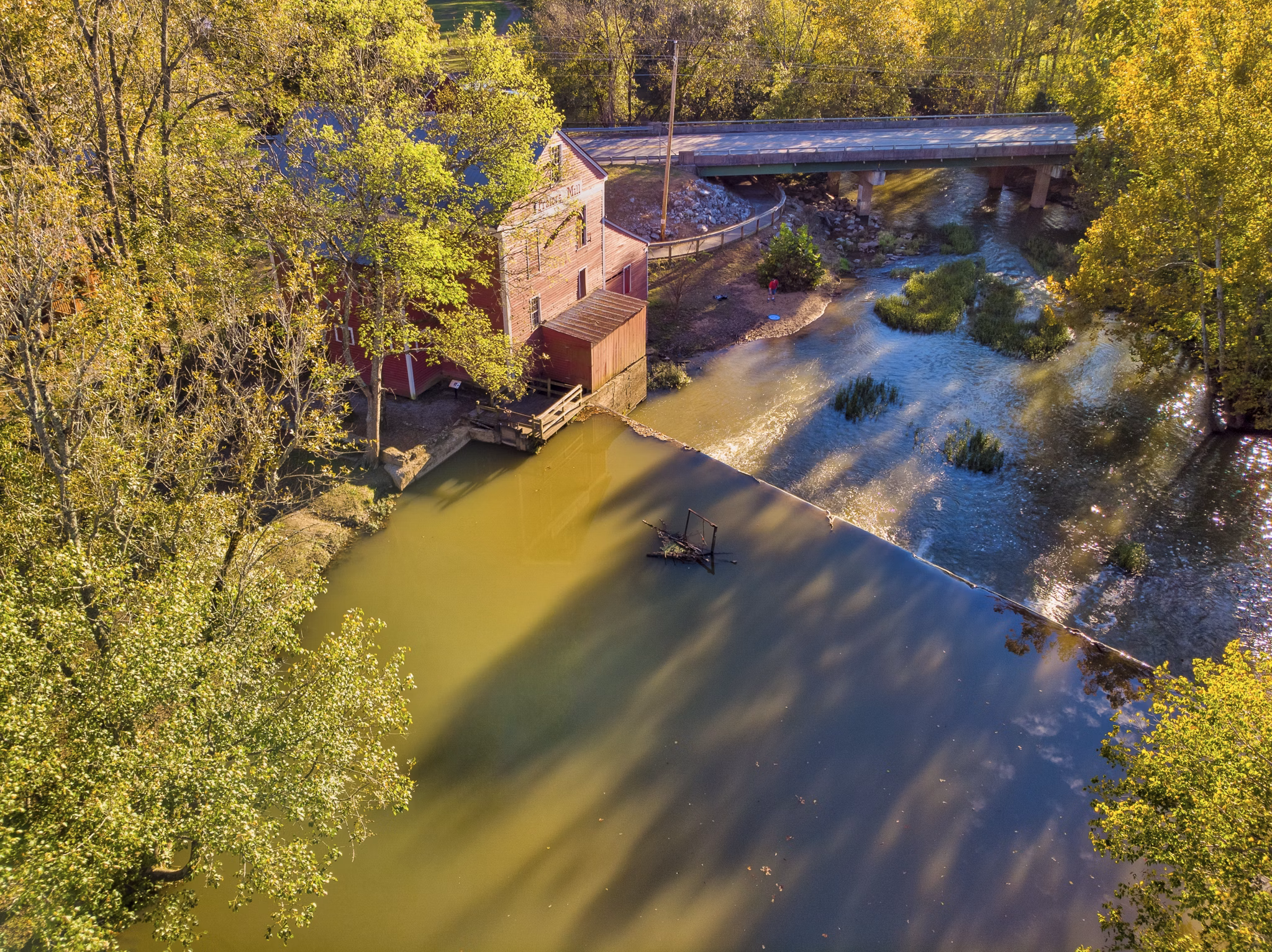 View of historic mill building beside creek in Dalton, GA with small dam creating a pond, surrounded by autumn trees.